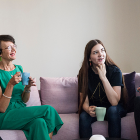 Neljä naista ja yksi mies istuvat sohvalla keskustelemassa. He hymyilevät ja heillä on kahvikupit kädessään.